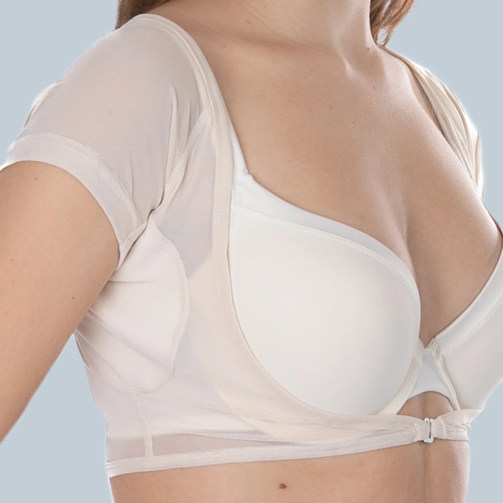 Studio foto vooraanzicht van vrouwelijk model die een ConfidenceForAll DryDress top draagt , een doorschijnend crème top met korte mouwen en zweetabsorberende, anti-transpirant zweetpads over een witte bh met het kledingstuk aan de voorkant dichtgemaakt met een haakje.