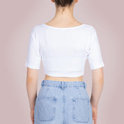 Studiofotografie van de achterkant van een vrouwelijk model dat een ConfidenceForAll Sweatproof witte crop top met korte mouwen en zweetpads die behandeld zijn met zilverionen draagt en een lichtblauwe spijkerbroek, staat met haar handen in haar zij tegen een lichtroze achtergrond.