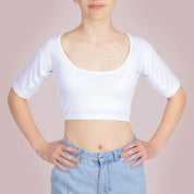 Studiofotografie van de voorkant van een vrouwelijk model dat een ConfidenceForAll Sweatproof witte crop top met korte mouwen en zweetpads die behandeld zijn met zilverionen draagt en een lichtblauwe spijkerbroek, staat met haar handen in haar zij tegen een lichtroze achtergrond.