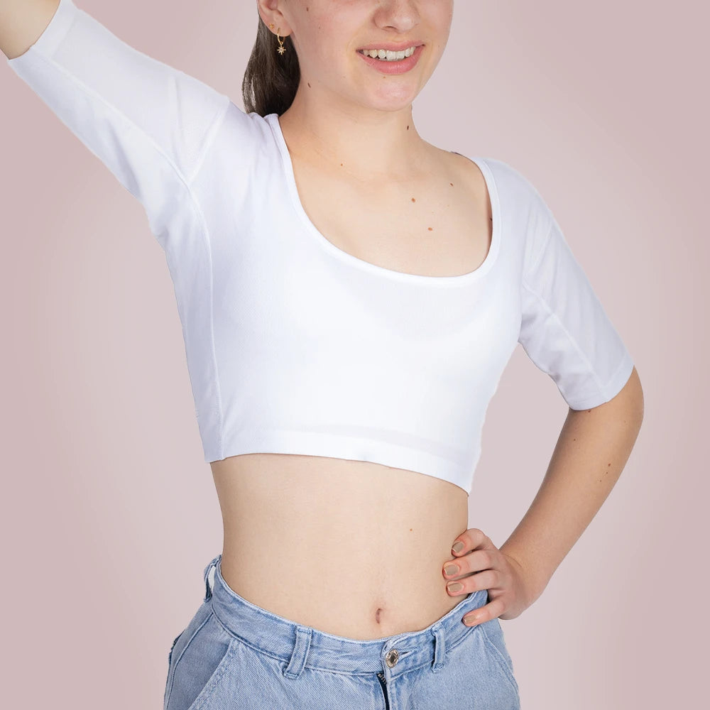 Een vrouwelijk model draagt een ConfidenceForAll Sweatproof witte crop top met korte mouwen en zweetpads die behandeld zijn met zilverionen, en een lichtblauwe jeans. Ze staat met één hand op haar heup en de andere arm omhoog om te laten zien waar de pad van haar shirt zit. Ze staat tegen een lichtroze achtergrond.