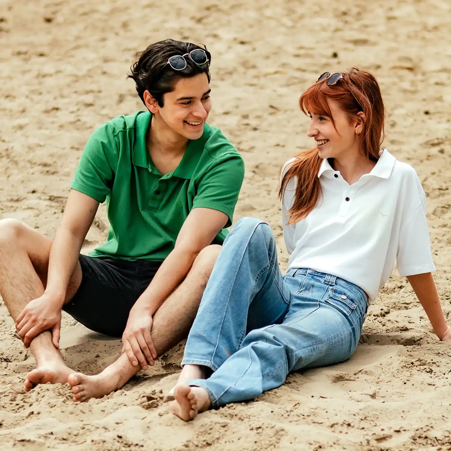 A young couple in casual summer outfits, a ConfidenceForAll green polo and a white polo, sitting on the beach, smiling and enjoying their time together