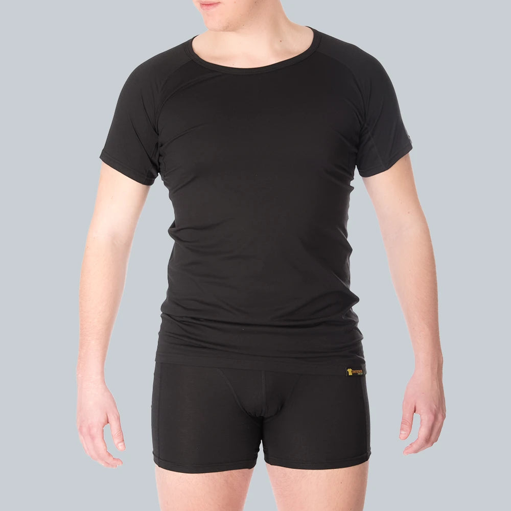 A male model wearing a fitted ConfidenceForAll black short-sleeve anti-transpirant sweatproof shirt and black ConfidenceForAll boxer briefs with sweat pads. Person stands against a plain grey blue background.
