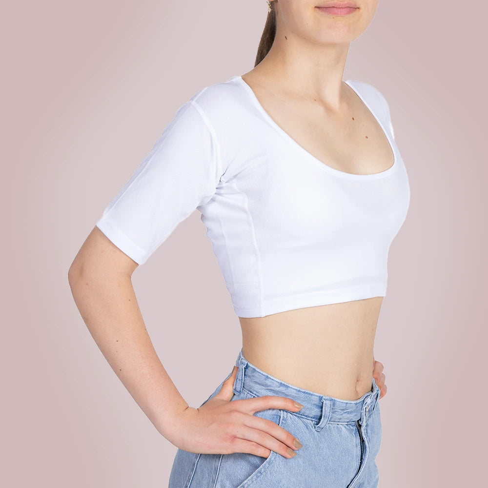 Een meisje draagt ​​een ConfidenceForAll Sweatproof witte crop top met korte mouwen en zweetpads die behandeld zijn met zilverionen en een lichtblauwe spijkerbroek. Ze staat met haar handen in haar zij tegen een lichtroze achtergrond.