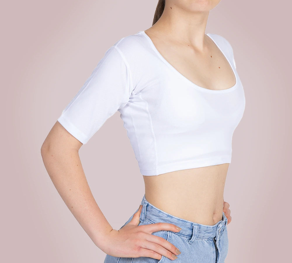 Een meisje draagt ​​een ConfidenceForAll Sweatproof witte crop top met korte mouwen en zweetpads die behandeld zijn met zilverionen en een lichtblauwe spijkerbroek. Ze staat met haar handen in haar zij tegen een lichtroze achtergrond.