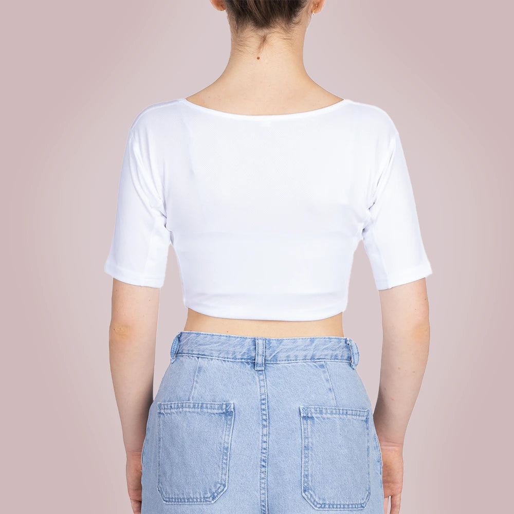 Studiofotografie van de achterkant van een vrouwelijk model dat een ConfidenceForAll Sweatproof witte crop top met korte mouwen en zweetpads die behandeld zijn met zilverionen draagt ​​en een lichtblauwe spijkerbroek, staat met haar handen in haar zij tegen een lichtroze achtergrond.