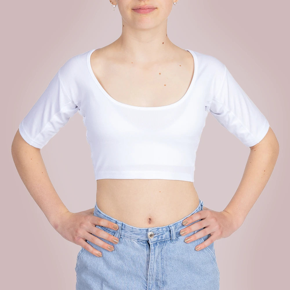 Studiofotografie van de voorkant van een vrouwelijk model dat een ConfidenceForAll Sweatproof witte crop top met korte mouwen en zweetpads die behandeld zijn met zilverionen draagt ​​en een lichtblauwe spijkerbroek, staat met haar handen in haar zij tegen een lichtroze achtergrond.
