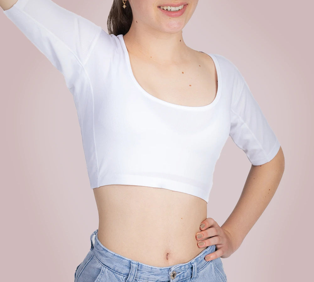 Een vrouwelijk model draagt ​​een ConfidenceForAll Sweatproof witte crop top met korte mouwen en zweetpads die behandeld zijn met zilverionen, en een lichtblauwe jeans. Ze staat met één hand op haar heup en de andere arm omhoog om te laten zien waar de pad van haar shirt zit. Ze staat tegen een lichtroze achtergrond.